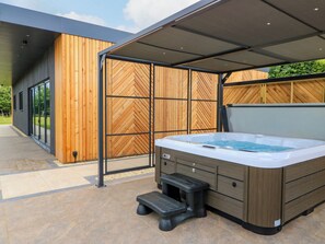 A hot tub with a wooden exterior, a covered patio area, and a modern building with large windows.