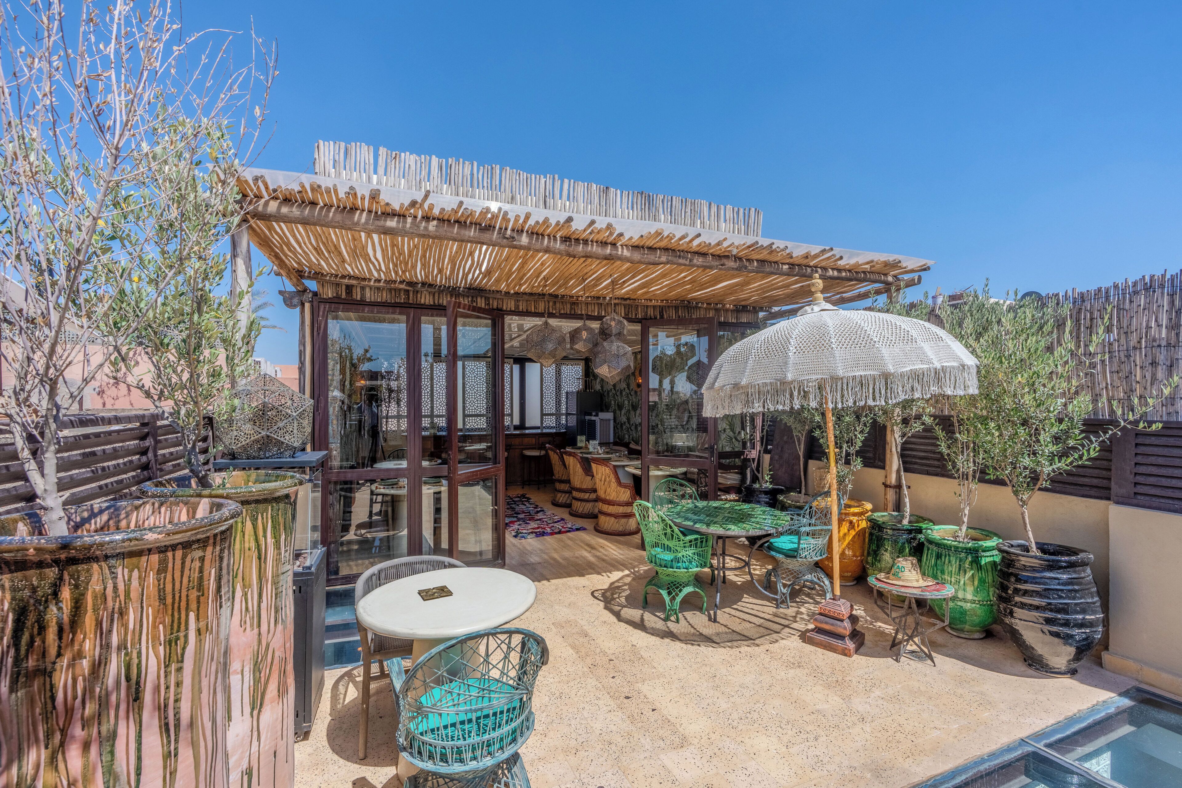 Un espace extérieur abrité, doté de tables et de chaises, d’une structure en verre et de plantes en pot.