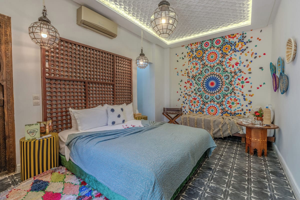 A bedroom with a bed, a wooden headboard, a colorful wall tapestry, and a small wooden table.