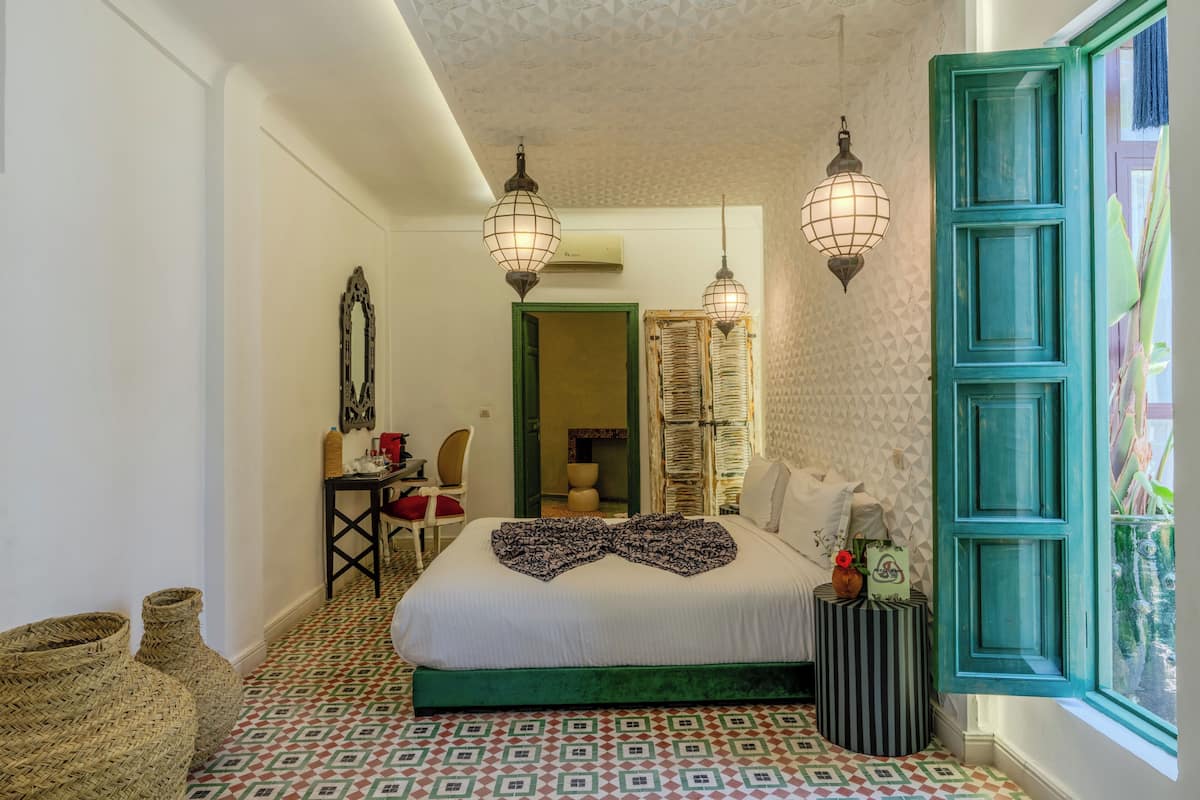 A bedroom with a bed, two hanging lamps, a window with teal shutters, and a patterned floor.