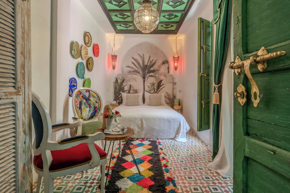 A narrow hallway leading to a bedroom with a bed, a chair, a table with a vase, and a colorful rug.