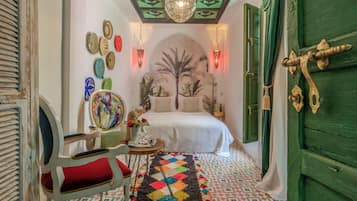 A narrow hallway leading to a bedroom with a bed, a chair, a table with a vase, and a colorful rug.