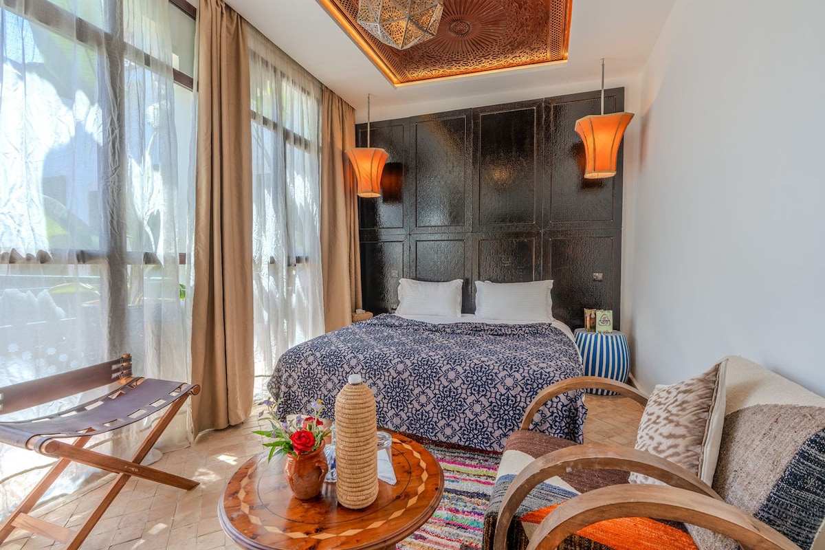 A hotel room with a large bed, a sofa, a small table with a vase and a candle, and a view of the outdoors.