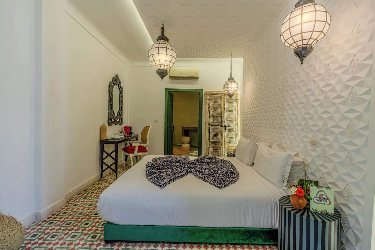 A hotel room with a bed, two hanging lamps, a mirror, a small table, and a chair.