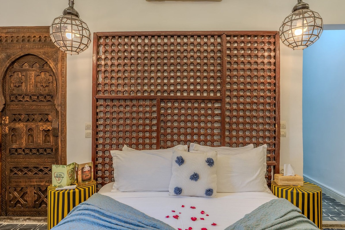 A room with a bed, a decorative wooden headboard, two bedside lamps, and a patterned wall.
