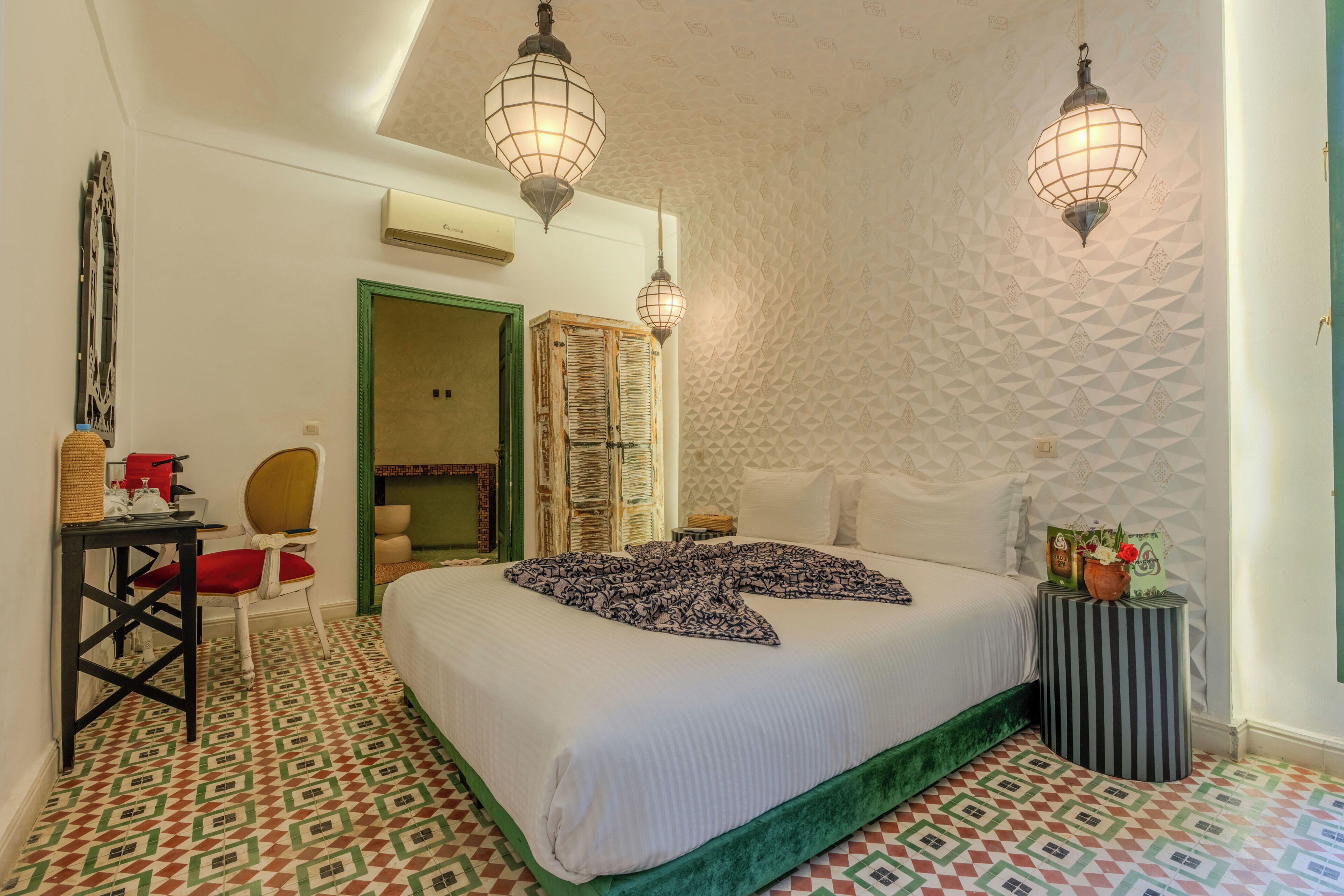 A hotel room with a bed, two standing lamps, a chair, a table, and a patterned floor.