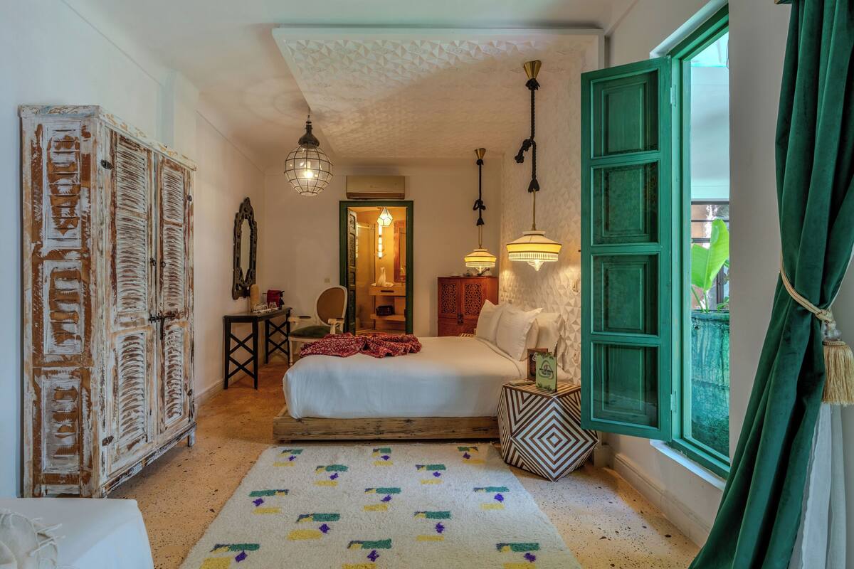 A bedroom with a bed, a wardrobe, a mirror, a bedside table, a lamp, and a window with curtains.