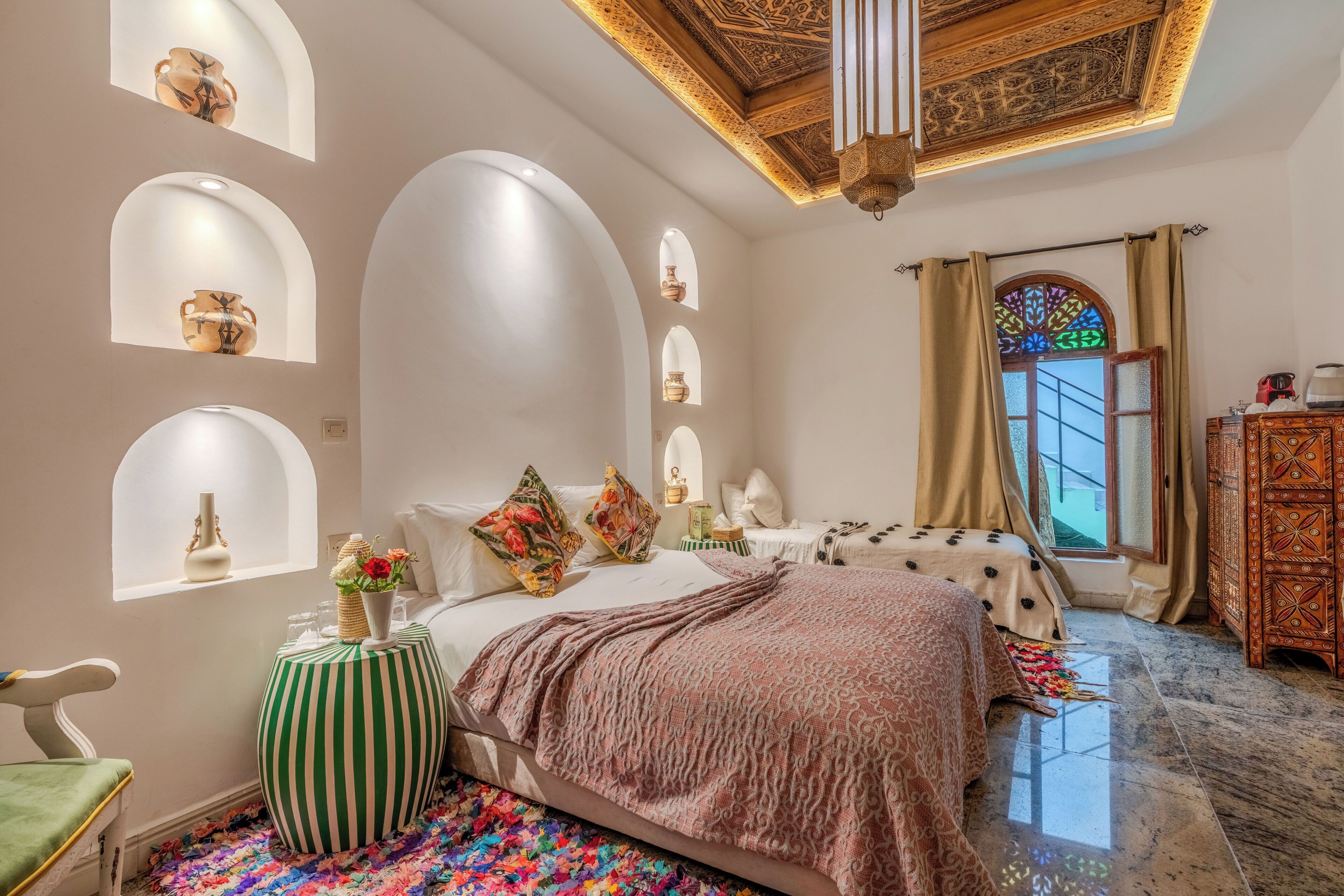 A bedroom with a bed, a green-striped ottoman, a patterned throw blanket, and a colorful rug.