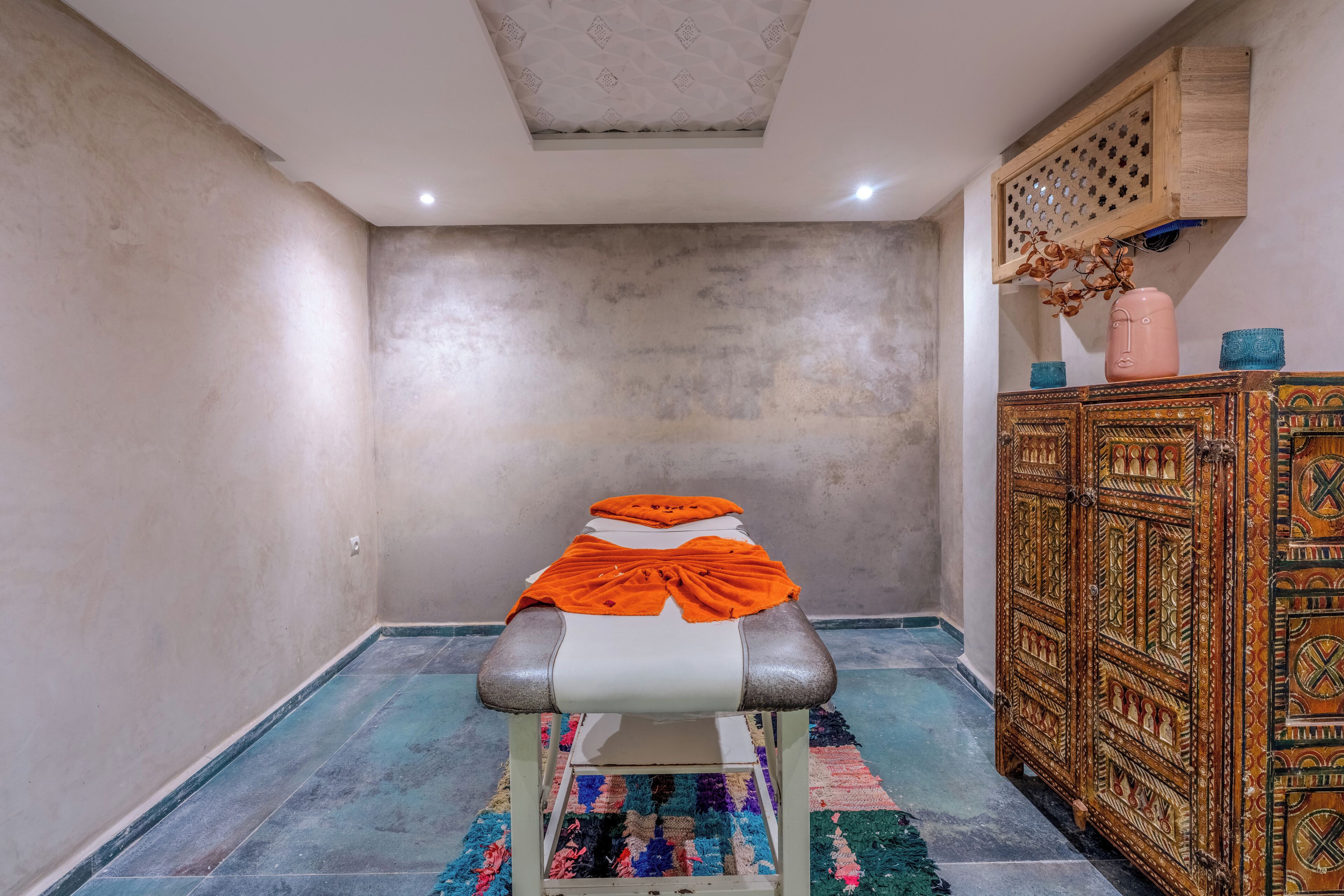 Una mesa de masaje con una toalla naranja, un tapete estampado y un mueble de madera con detalles decorativos.