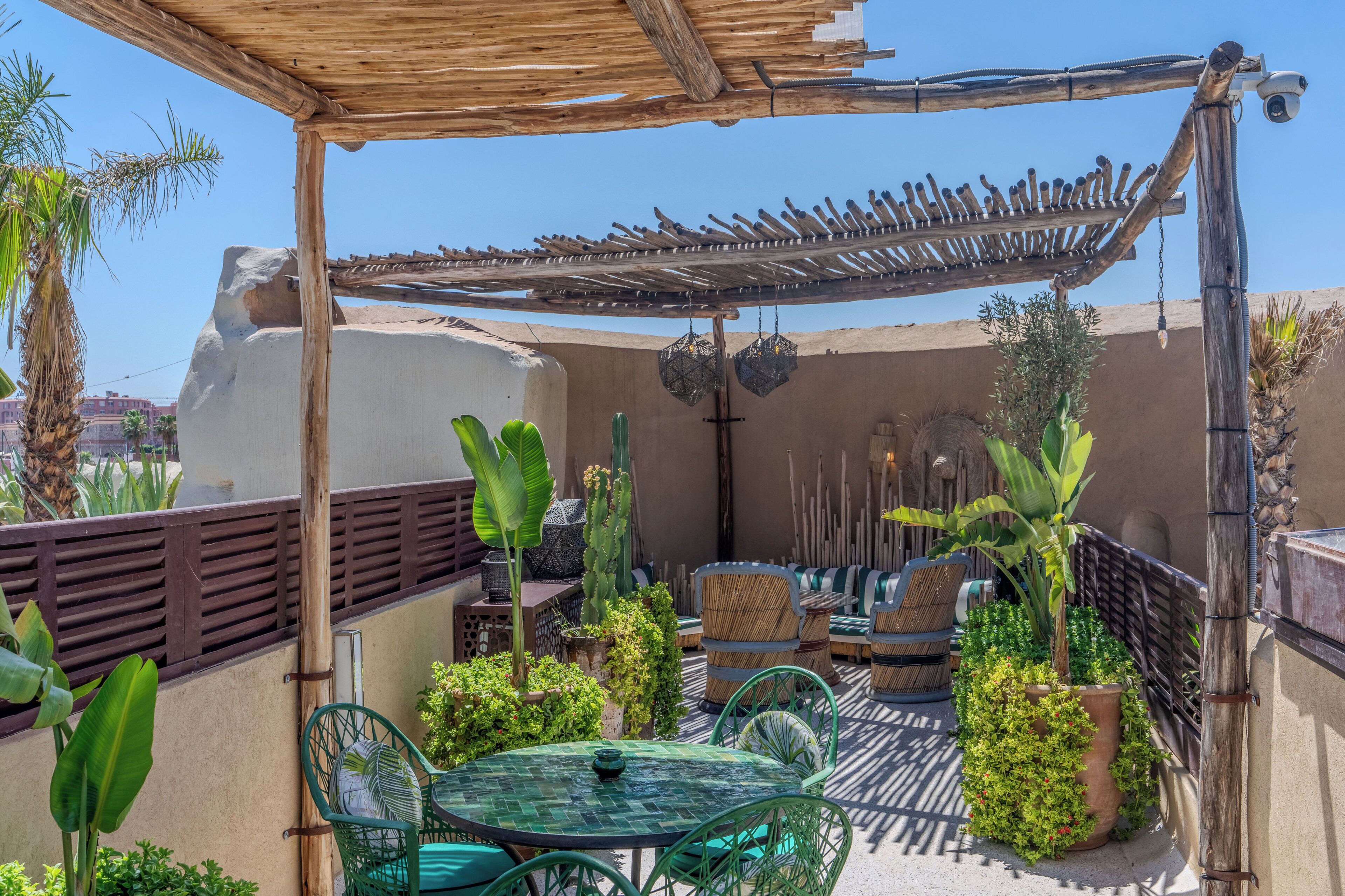 Un espace extérieur abrité, aménagé avec une table et des chaises, entouré de plantes et surmonté d’une pergola en bois.