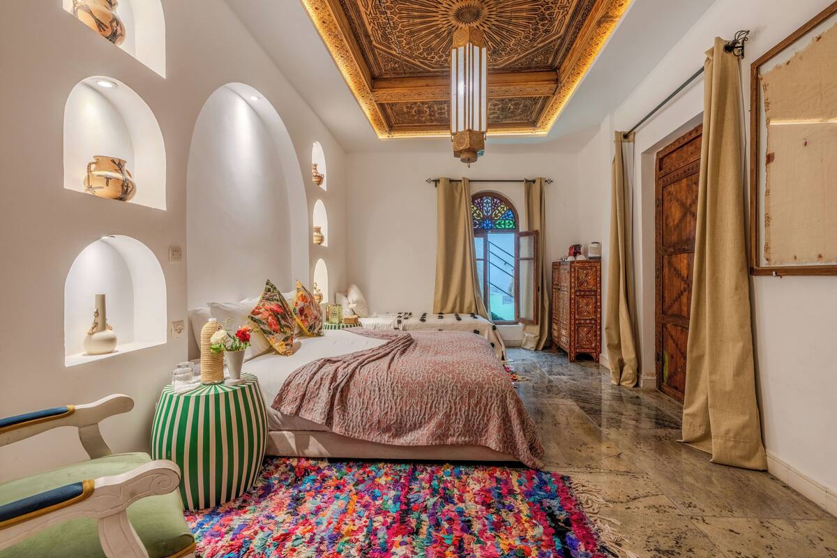 A bedroom with a bed, a green-striped ottoman, a colorful rug, a wooden cabinet, and a decorative ceiling.