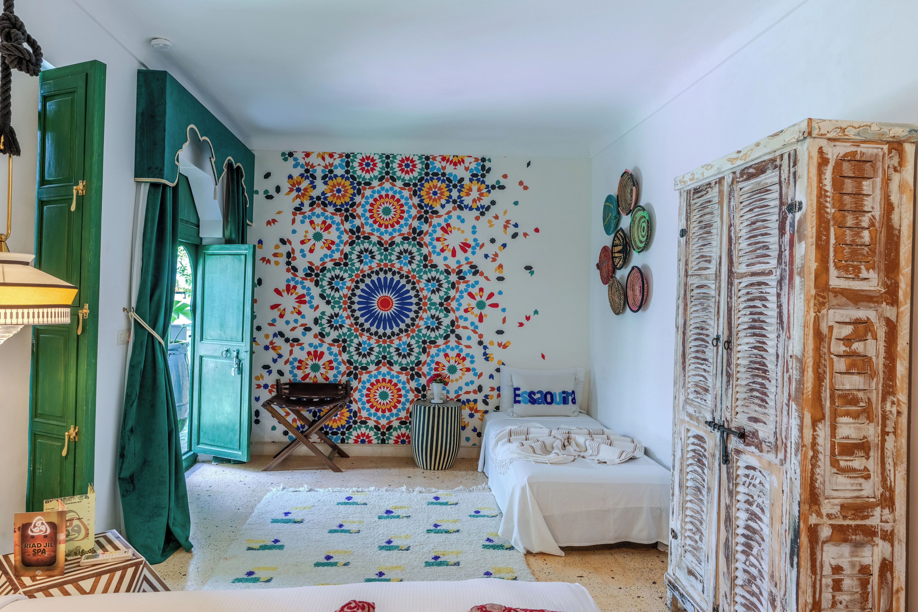 A room with a bed, a green door, a colorful wall tapestry, and a small table with a lamp.