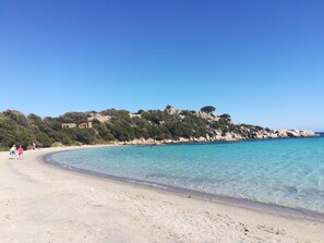 Ein Strand mit klarem türkisblauem Wasser, einem Sandufer und felsigen Klippen mit Vegetation.