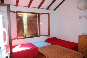Ein Zimmer mit einem Holztisch, zwei Einzelbetten mit roter Bettwäsche, einem Holzschrank und einem Fenster mit geöffneten Fensterläden.