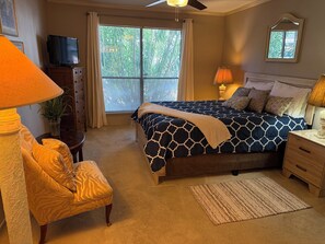 Een slaapkamer met een bed, een stoel, een bureau, een lamp, een televisie en een raam met jaloezieën.