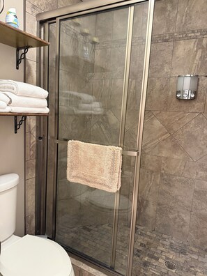 Een douche met een glazen deur, een handdoekrek met opgevouwen handdoeken en een zeepdispenser aan de muur.