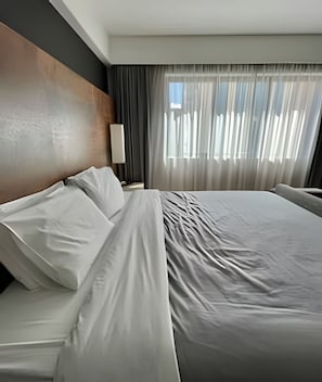 Une chambre d’hôtel avec un grand lit, une table de chevet, une lampe et une fenêtre avec des rideaux légers.