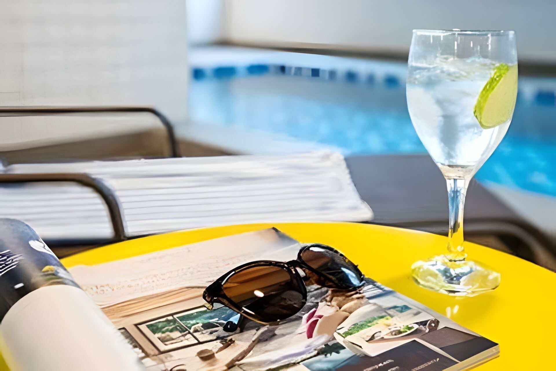Escena junto a la piscina con un vaso de agua, gafas de sol y una revista sobre una bandeja amarilla.