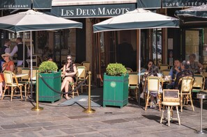 Outdoor seating with tables and chairs, a green planter, and a sign for 'Les Deux Magots.' - 3 bedroom apartment (Paris)