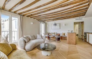 A modern living room with a curved sofa, a glass coffee table, and a dining area with metal chairs. - LIVE LIKE PARISIAN IN THE MOST FAMOUS NEIGHBOURHOOD-SAINT GERMAIN - RUE DAUPHINE (Paris)