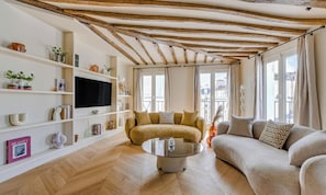 A modern living room with a sofa, a coffee table, and a TV. There are shelves with decorative items and a window with curtains. - A REFINED PARISIAN ELEGANCE WITH CONTEMPORARY COMFORT-RUE DAUPHINE SAINT GERMAIN (Paris)