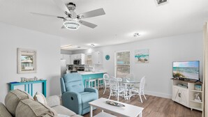 A living room with a sofa, a blue armchair, a dining table with chairs, a kitchen area, and a TV. - Beachy 2BR Escape with Pool Views  | St. Martin Beachwalk Villa 314 (Destin)