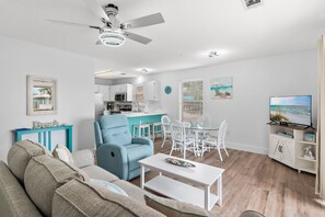 A living room with a sofa, a blue armchair, a dining table with chairs, a kitchen area, and a TV. - Beachy 2BR Escape with Pool Views  | St. Martin Beachwalk Villa 314 (Destin)