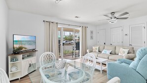 A living room with a glass table, white chairs, a beige sofa, and a television on a cabinet. - Beachy 2BR Escape with Pool Views  | St. Martin Beachwalk Villa 314 (Destin)