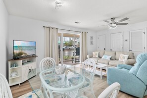 A living room with a glass table, white chairs, a beige sofa, and a television on a cabinet. - Beachy 2BR Escape with Pool Views  | St. Martin Beachwalk Villa 314 (Destin)