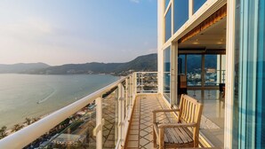 Balcón con vista al mar y colinas distantes, un banco de madera y una puerta de cristal que da a un espacio interior.