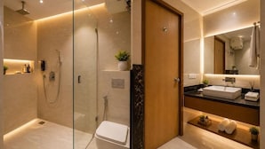 A modern bathroom with a glass-enclosed shower, a wooden door, a white toilet, a sink with a black countertop, and a mirror. - Hotel The Capital Stay (New Delhi)