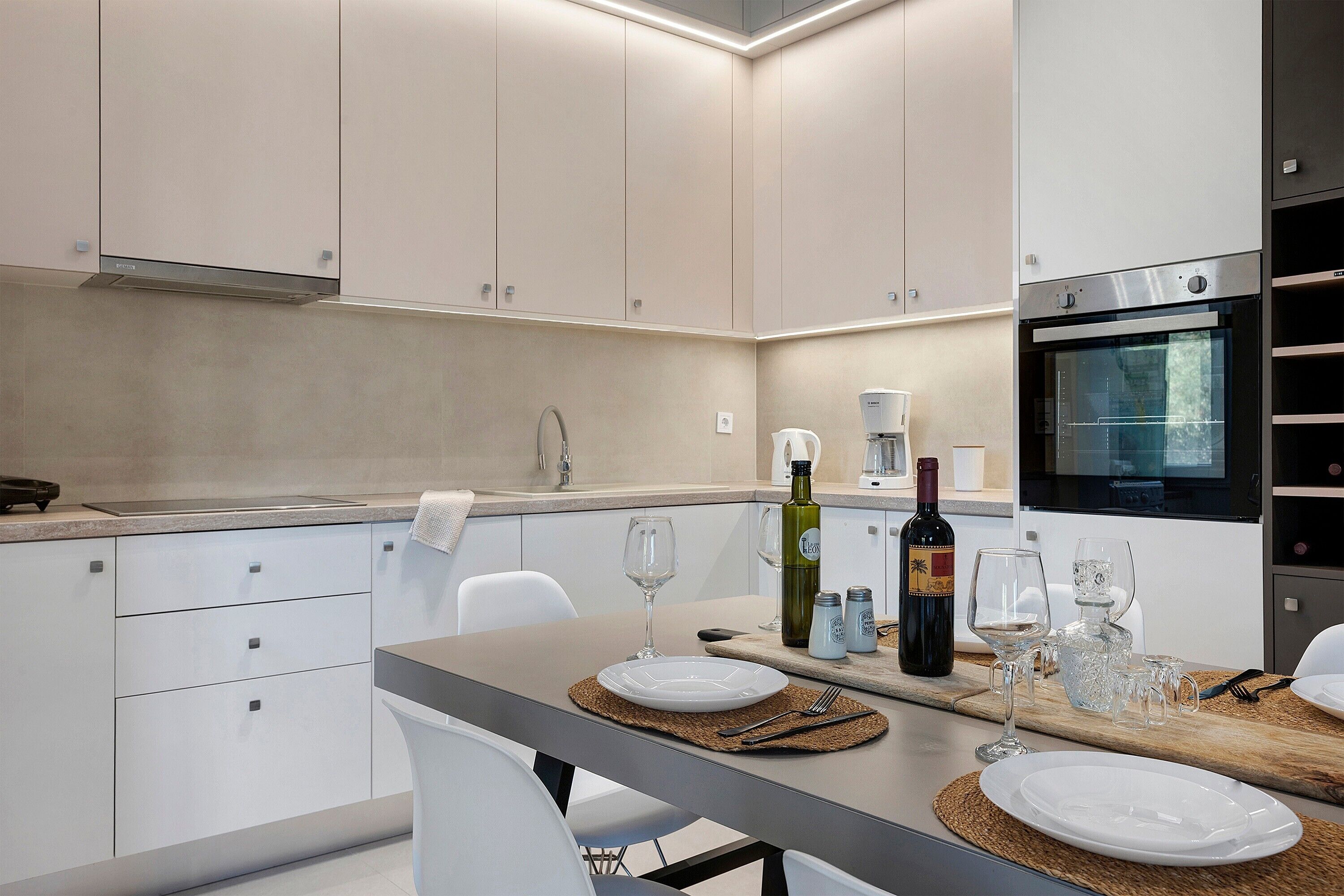 Een moderne keuken met witte kasten, een ingebouwde oven en een eethoek met borden, glazen en een fles.