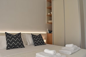Cama bem arrumada com duas almofadas, um conjunto de toalhas dobradas e uma prateleira de madeira com artigos de higiene pessoal.