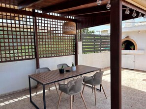 Eine überdachte Terrasse mit Tisch, Stühlen, einem gewebten Korb und einem Backofen im Hintergrund.