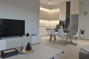 Cozinha moderna com armários brancos, área de jantar com cadeiras brancas e uma televisão de tela plana sobre um console branco.