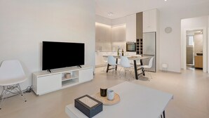 Un salon moderne doté d’un téléviseur à écran plat, d’un coin repas avec des chaises, d’une cuisine avec des placards et d’une horloge murale.