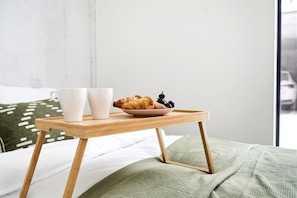 Ein Bett mit grüner Decke, ein Nachttisch aus Holz mit einer Schale, die ein Croissant, zwei weiße Tassen und eine Schüssel mit Beeren enthält.