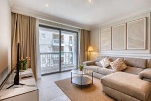 Ein modernes Wohnzimmer mit einer Couch, einem Couchtisch und einem Fernseher. Ein Balkon bietet Blick auf umliegende Gebäude.