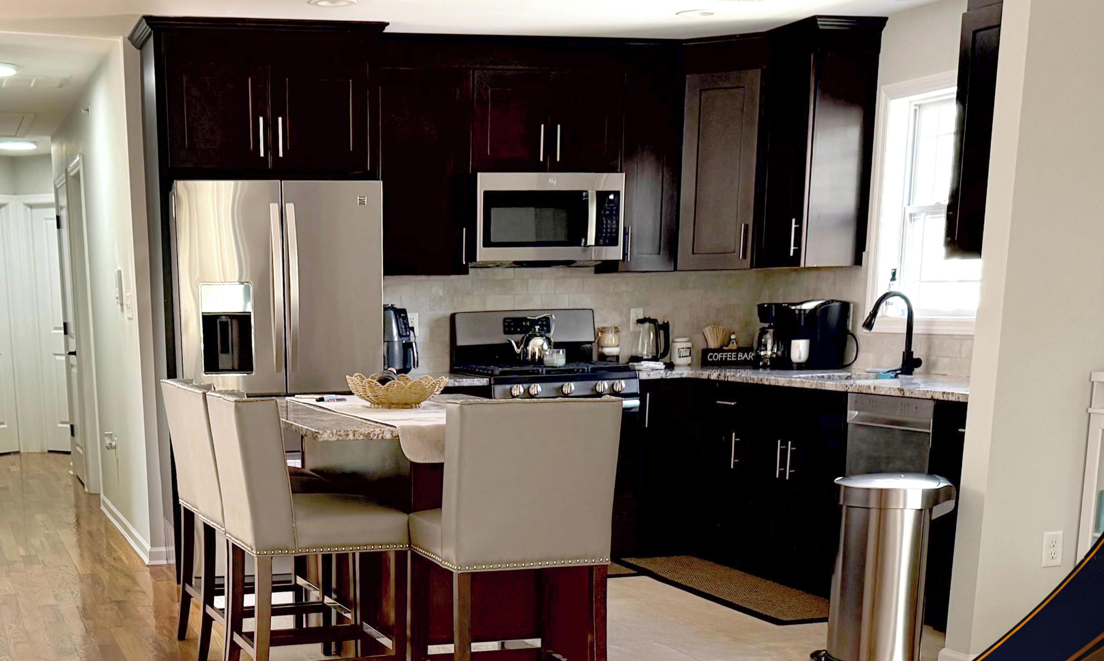 A modern kitchen with dark cabinetry, stainless steel appliances, a coffee station, and a bar seating area.. Modern 3BR Home Near NYC, Newark Airport & MetLife Stadium