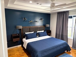 Un dormitorio con una cama grande, dos mesitas de noche, un ventilador de techo y una ventana con cortinas.