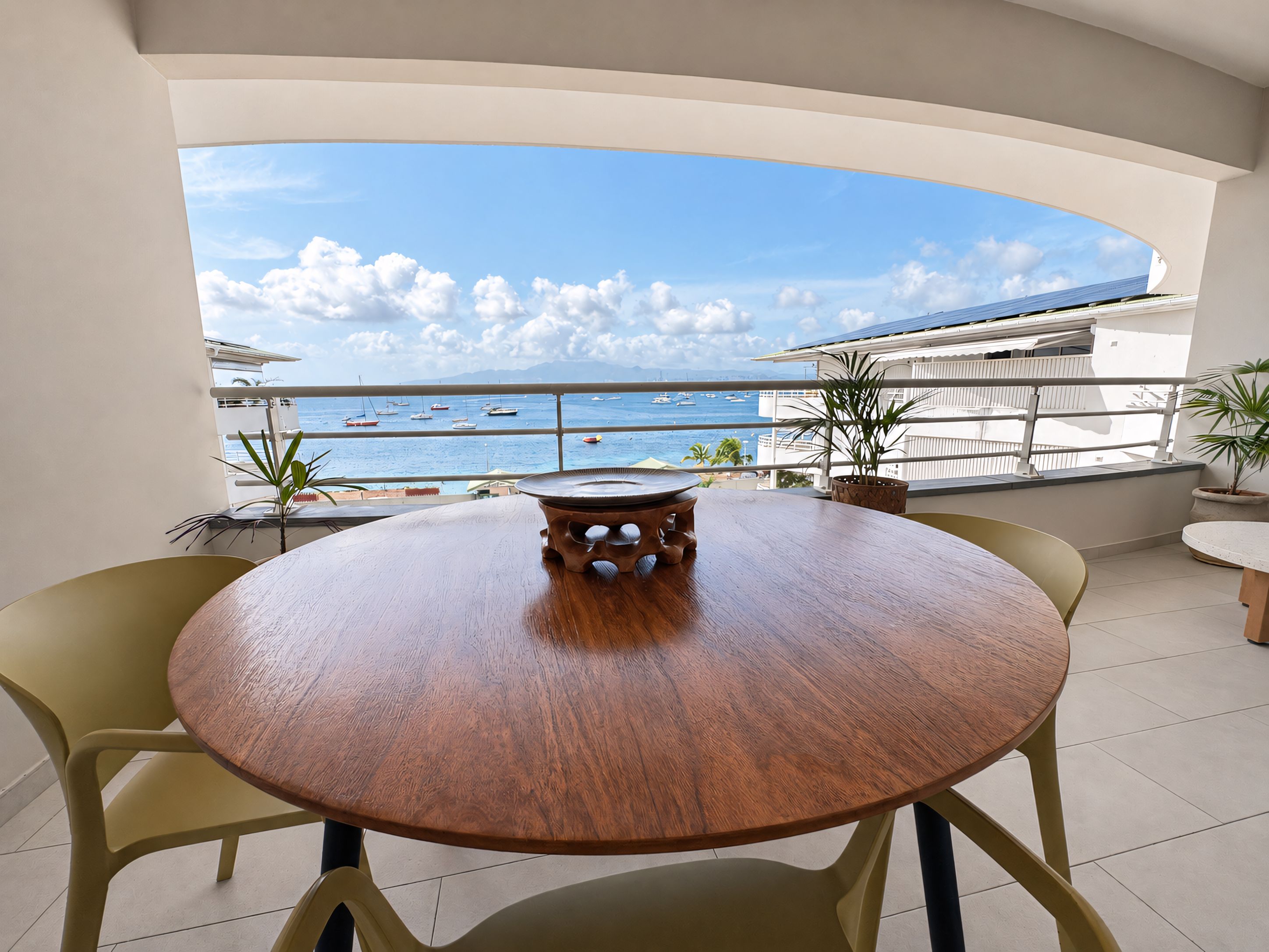 A round wooden table with chairs on a balcony overlooking the sea with boats.