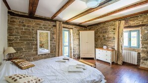 A bedroom with stone walls, a wooden ceiling, a bed with white linens, a dresser, and a window with curtains. - Casa Biloslavi with pool (Groznjan)