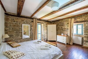 Ein Schlafzimmer mit Steinwänden, Holzbalkendecke, einem Bett mit weißen Leinentüchern, einem Schminktisch und einem Fenster mit Vorhängen.