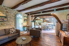 Ein rustikales Interieur mit Steinwänden, Holzbalken, einem Kamin aus Stein, einem Essbereich und einer durch einen Bogen sichtbaren Küche.