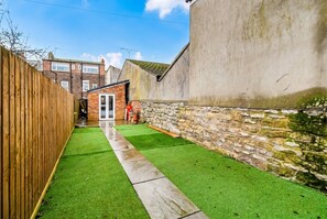 A narrow backyard with artificial grass, a wooden fence, a stone wall, and a small brick structure. - Cosy Tiny Home for 2 with Garden · Walk to Filey Beach (North Yorkshire)
