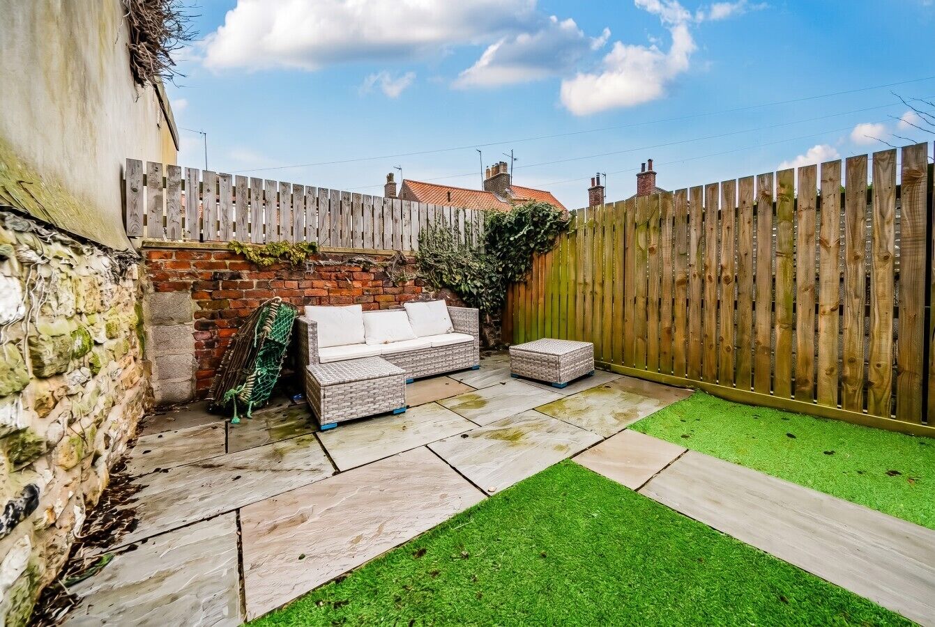 A backyard with a stone wall, a wooden fence, a brick wall, a green lawn, and a white outdoor sofa.