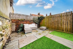 A backyard with a stone wall, a wooden fence, a brick wall, a green lawn, and a white outdoor sofa. - Cosy Tiny Home for 2 with Garden · Walk to Filey Beach (North Yorkshire)