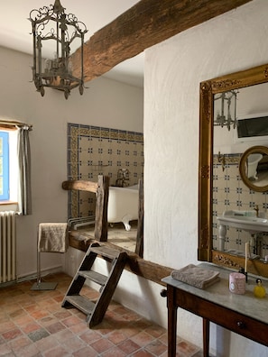 Un baño rústico con una escalera de madera, un candelabro antiguo, un espejo grande con marco dorado y una pared de azulejos estampados.