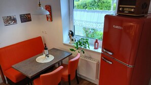A compact kitchen with a red refrigerator, a dining table with two chairs, a small window with a view of greenery, and a wall clock. - Ground-Level Holiday Home by the Salt Marshes (Sande)