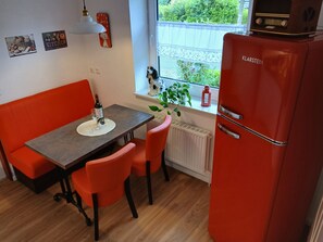 Eine kleine Küche mit einem roten Kühlschrank, einem Esstisch mit zwei Stühlen, einem kleinen Fenster mit Blick auf Grünflächen und einer Wanduhr.